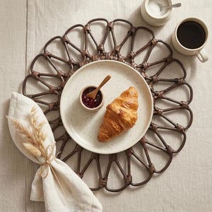 Könnte beinhalten: Ein Frühstücksarrangement mit einem braunen, gewebten Platzset, einem weißen Teller mit Croissant und Marmelade, einem kleinen Holzlöffel, einer Serviette mit getrocknetem Weizen, einer Kaffeetasse und einer Zuckerdose.