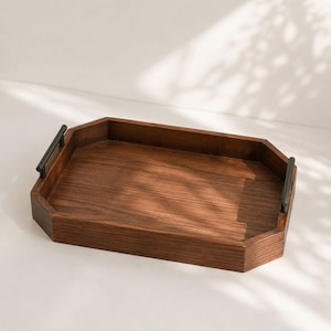 Puede incluir: Una bandeja de servicio rectangular de madera con esquinas biseladas y un acabado de veta de madera marrón intenso. La bandeja cuenta con dos asas delgadas de metal negro mate fijadas en los lados cortos, facilitando su transporte.