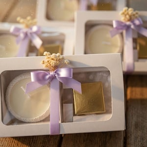 Puede incluir: Una caja de regalo rectangular blanca con ventana transparente que contiene una vela redonda blanca en un soporte texturizado y un pequeño objeto cuadrado de color dorado. La caja está decorada con un lazo de satén lila y flores secas.