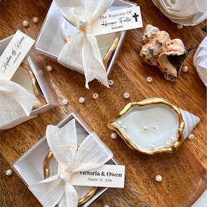 May include: Square clear gift boxes containing gold-rimmed shell-shaped candles, each tied with a cream-colored ribbon, a pearl accent, and a personalized tag. One tag reads The Baptism Of Harper Foster 09.09.2027, another Sophia & Lucas 30.05.2026, and another Victoria & Owen March 07, 2026.