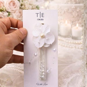 May include: A white card with the initials "T|E" and the date "17/04/2026" printed in black. A white flower with a pearl center and small pearls is attached to the card. A small glass vial filled with pearls is also attached. The words "With Love" are printed at the bottom.