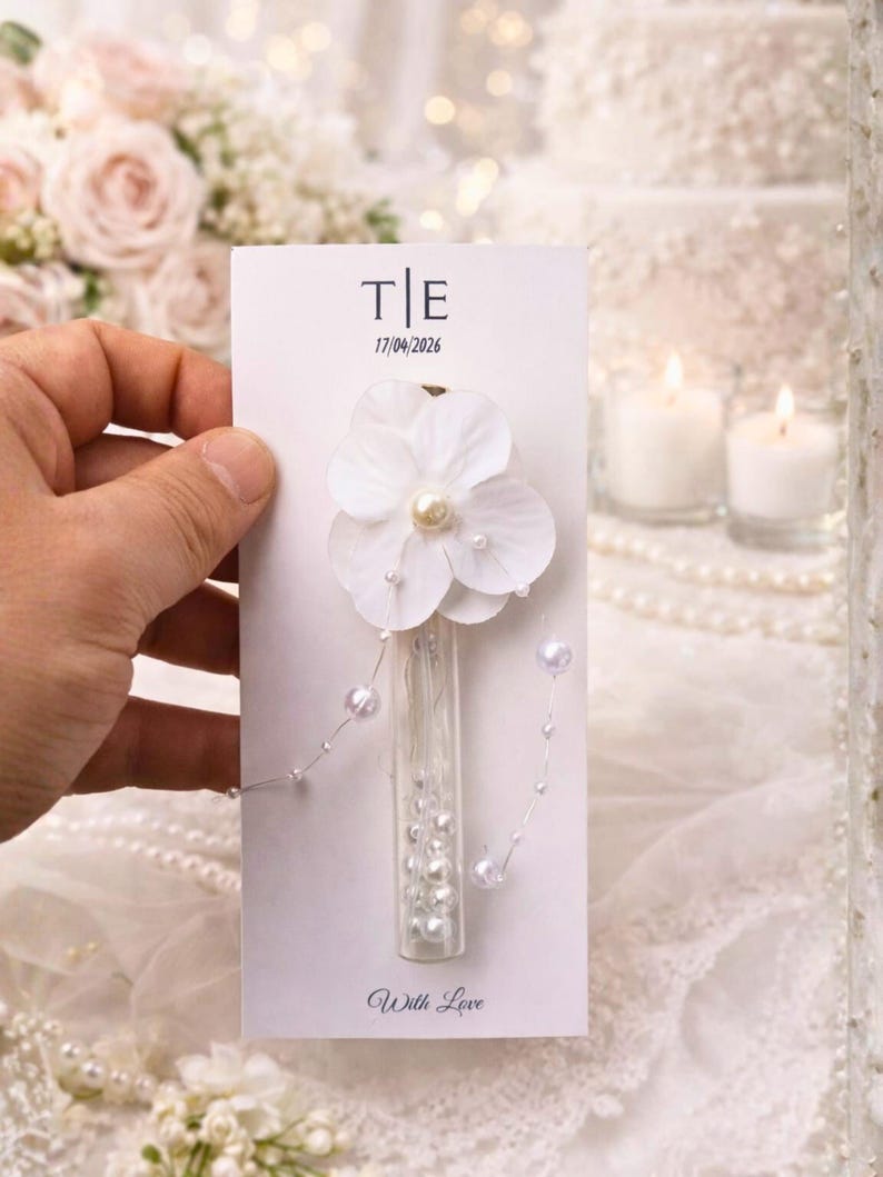 May include: A white card with the initials "T|E" and the date "17/04/2026" printed in black. A white flower with a pearl center and small pearls is attached to the card. A small glass vial filled with pearls is also attached. The words "With Love" are printed at the bottom.