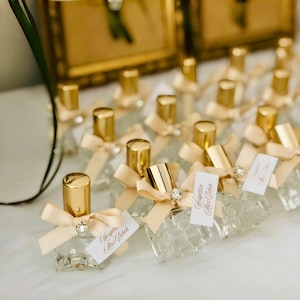 May include: A collection of small, clear glass perfume bottles with gold caps, each adorned with a beige ribbon bow, a small clear rhinestone, and a white rectangular tag featuring the printed text Hosgeldin Alaz Bebek in script font.