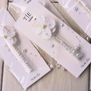 May include: White floral hair clips with pearl accents, packaged on white cards. Each card features the initials "T|E" and the date "17/04/2026". The words "With Love" are printed on the bottom of each card. The clips are in clear plastic.