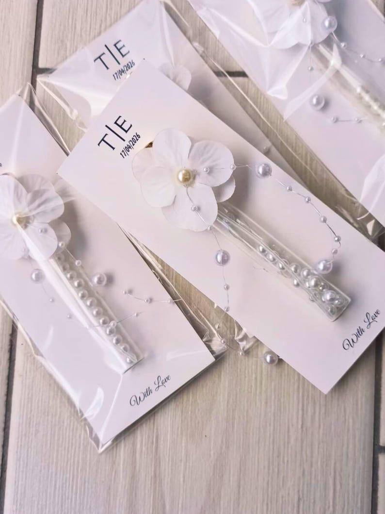 May include: White floral hair clips with pearl accents, packaged on white cards. Each card features the initials "T|E" and the date "17/04/2026". The words "With Love" are printed on the bottom of each card. The clips are in clear plastic.