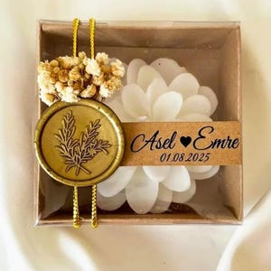 Puede incluir: Caja de regalo decorativa con una vela blanca en forma de flor. La caja está sellada con un sello de cera dorado y una ramita de flores secas. El texto "Asel Emre 01.08.2025" está impreso en una etiqueta marrón.