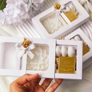 May include: A white rectangular gift box with a clear window containing a white flower-shaped candle, three white oval candies, and a square gold-colored chocolate bar with the letters ND and text printed on the front, tied with a white ribbon.