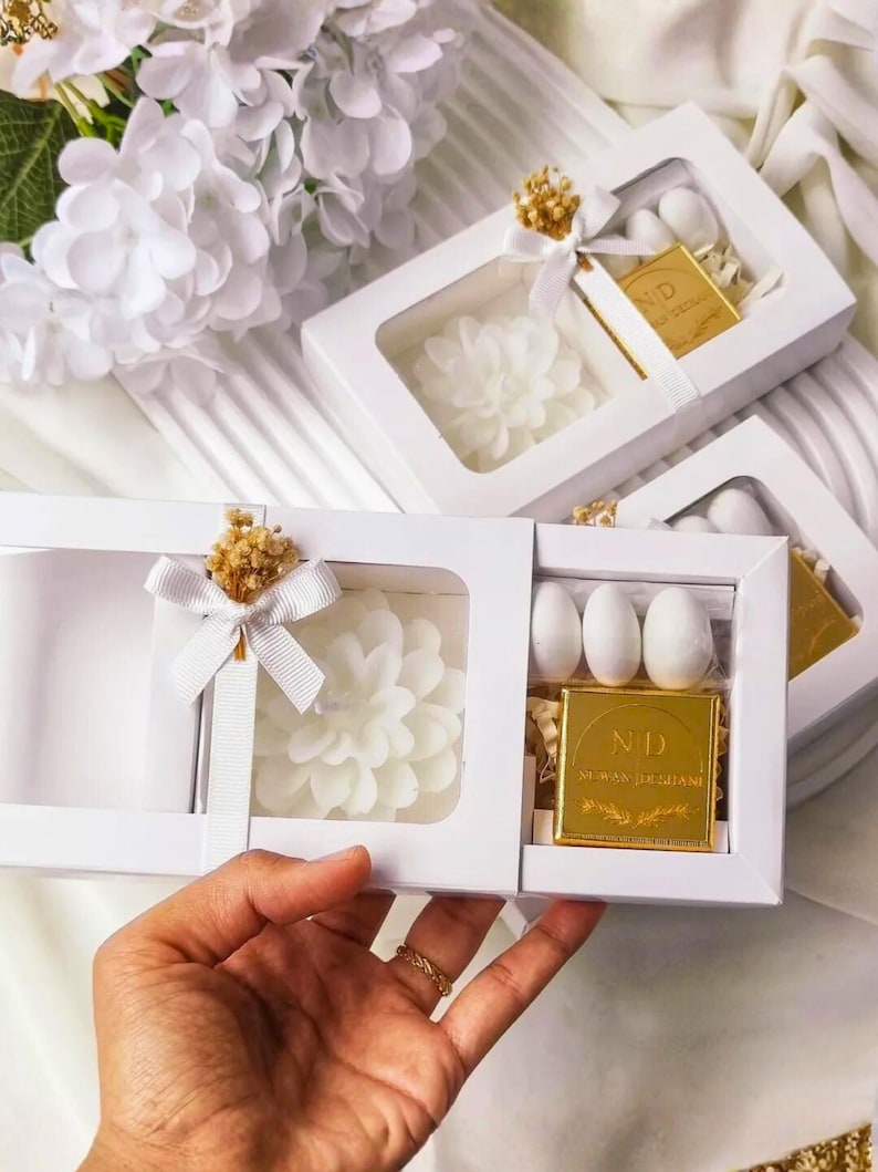 May include: A white rectangular gift box with a clear window containing a white flower-shaped candle, three white oval candies, and a square gold-colored chocolate bar with the letters ND and text printed on the front, tied with a white ribbon.