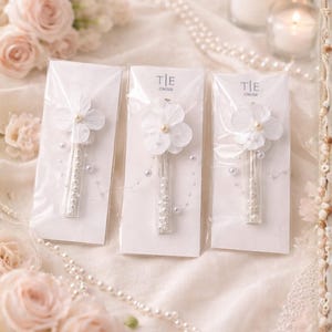 May include: Three white flower hair clips with pearl accents are displayed on white cards. Each clip features a delicate flower and a stem adorned with small pearls. The background includes roses, pearls, and candles, creating a soft, romantic aesthetic.