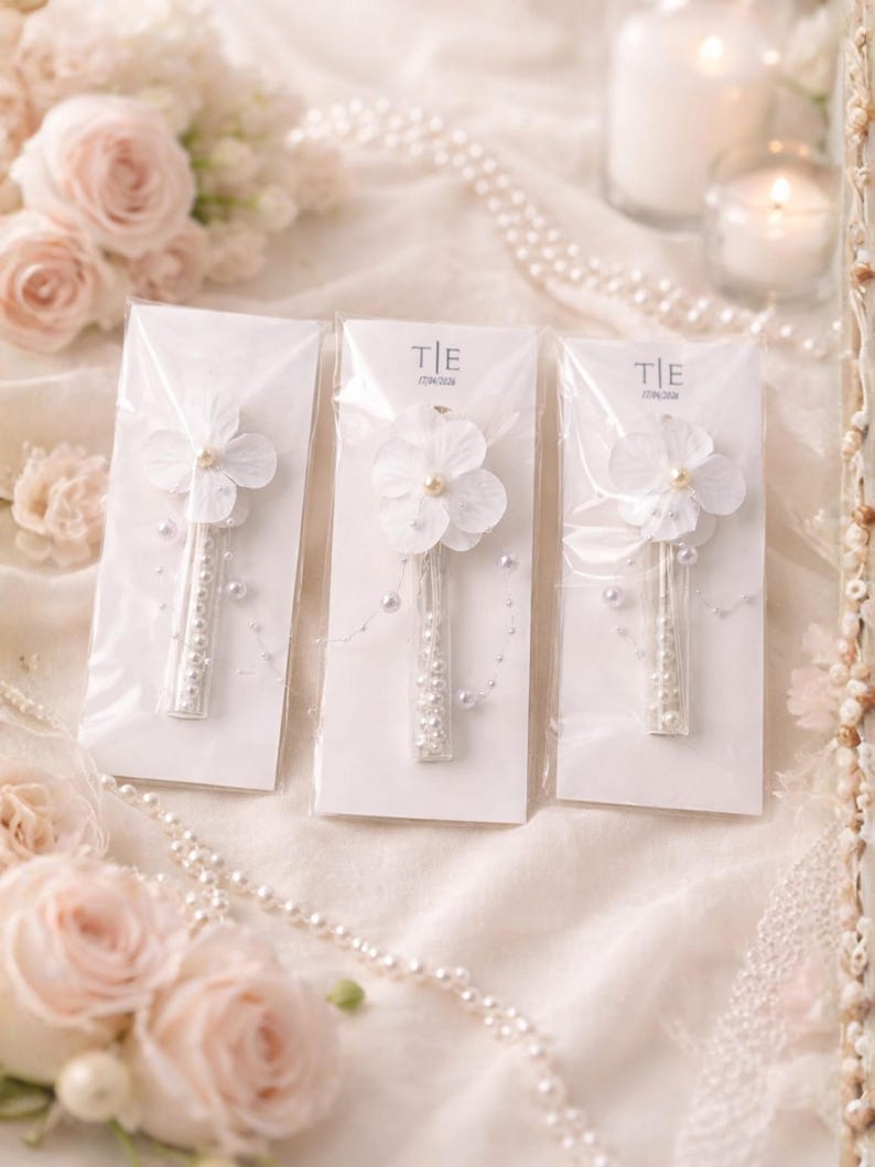 May include: Three white flower hair clips with pearl accents are displayed on white cards. Each clip features a delicate flower and a stem adorned with small pearls. The background includes roses, pearls, and candles, creating a soft, romantic aesthetic.