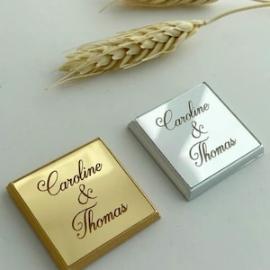 May include: Two square, flat items with a reflective finish, one in gold and one in silver. Each piece features the engraved text "Caroline & Thomas" in a cursive script font. Two dried wheat stalks are positioned above the square items.