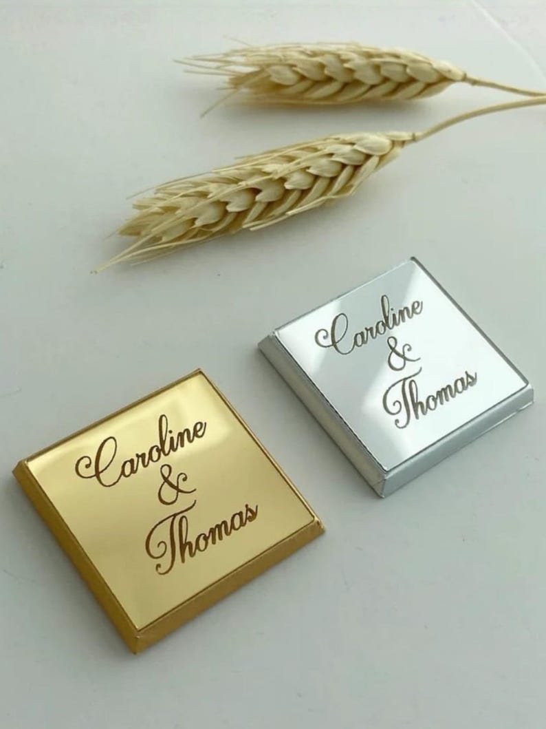May include: Two square, flat items with a reflective finish, one in gold and one in silver. Each piece features the engraved text "Caroline & Thomas" in a cursive script font. Two dried wheat stalks are positioned above the square items.