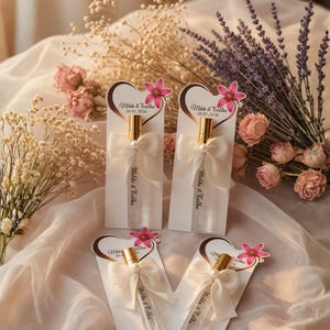 May include: Four perfume bottles with gold caps and white bows are displayed on a draped surface, each attached to a card with a heart design and the text "Melda & Falka 30.01.2026". Pink flowers and dried floral arrangements surround the bottles.