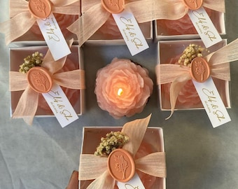 Op de afbeelding: Een verzameling lichtroze roosvormige kaarsen in vierkante witte doosjes. Elk doosje is versierd met een perzikkleurig waszegel, een transparant lint, een takje gedroogde bloemen en een wit label met de tekst Elif & Suka 26.10.2024 erop gedrukt.