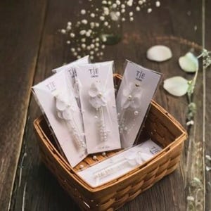 May include: A woven basket filled with several packaged tie clips. Each clip is white with a floral design and pearl accents. The packaging has the word "TIE" printed on it. The basket sits on a dark wooden surface.