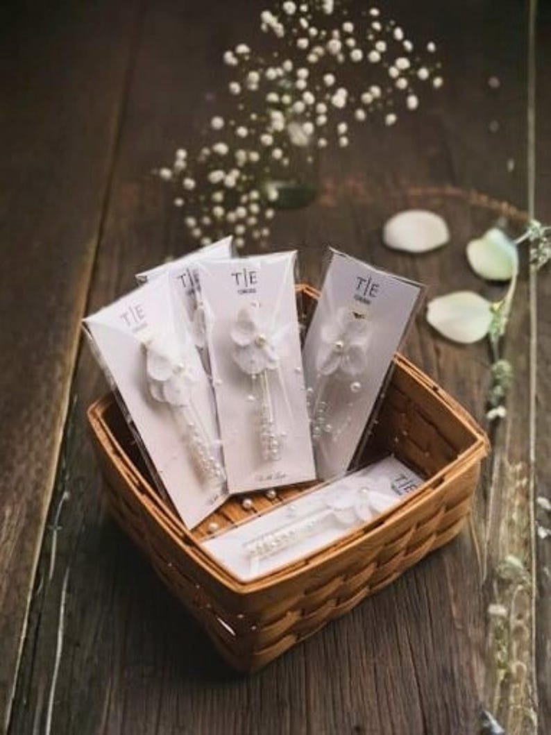 May include: A woven basket filled with several packaged tie clips. Each clip is white with a floral design and pearl accents. The packaging has the word "TIE" printed on it. The basket sits on a dark wooden surface.
