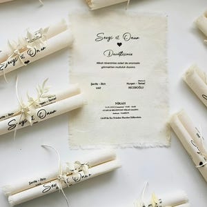 Könnte beinhalten: Cremefarbene Hochzeitseinladungen aus Stoff mit ausgefransten Rändern und schwarzem Aufdruck mit dem Text Sevgi & Onur sowie Veranstaltungsdetails. Jede Einladung ist gerollt und mit einer weißen Schnur und einem kleinen getrockneten Zweig verziert.