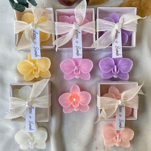 Op de afbeelding: Orchideevormige kaarsen in geel, roze, paars, wit en perzik. Sommige zitten in vierkante doosjes met crèmekleurige linten en labels met de tekst Arzu & Semih. Eén roze kaars brandt. De kaarsen hebben gedetailleerde bloemblaadjes en een lont.