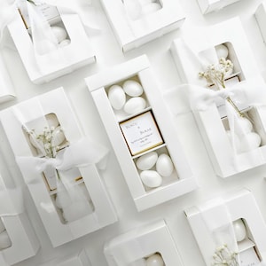 May include: A collection of rectangular white gift boxes with clear windows, each containing white almond candies and a central square card printed with the names Burcu & Burak and the date 01.06.2024, tied with a white ribbon and a sprig of dried baby's breath.