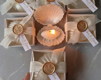 May include: Several small gift boxes tied with ivory ribbons and sealed with gold wax stamps. Each box contains a white item and a tag that reads "Inci & Emre". A seashell-shaped candle holder with a lit candle is also present.