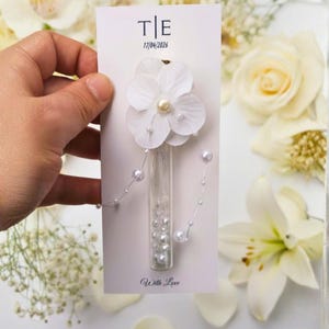 May include: A white floral wedding favor card with the initials "T|E" and the date "17/04/2026". The card features a white flower with a pearl center, a glass vial filled with pearls, and the words "With Love".