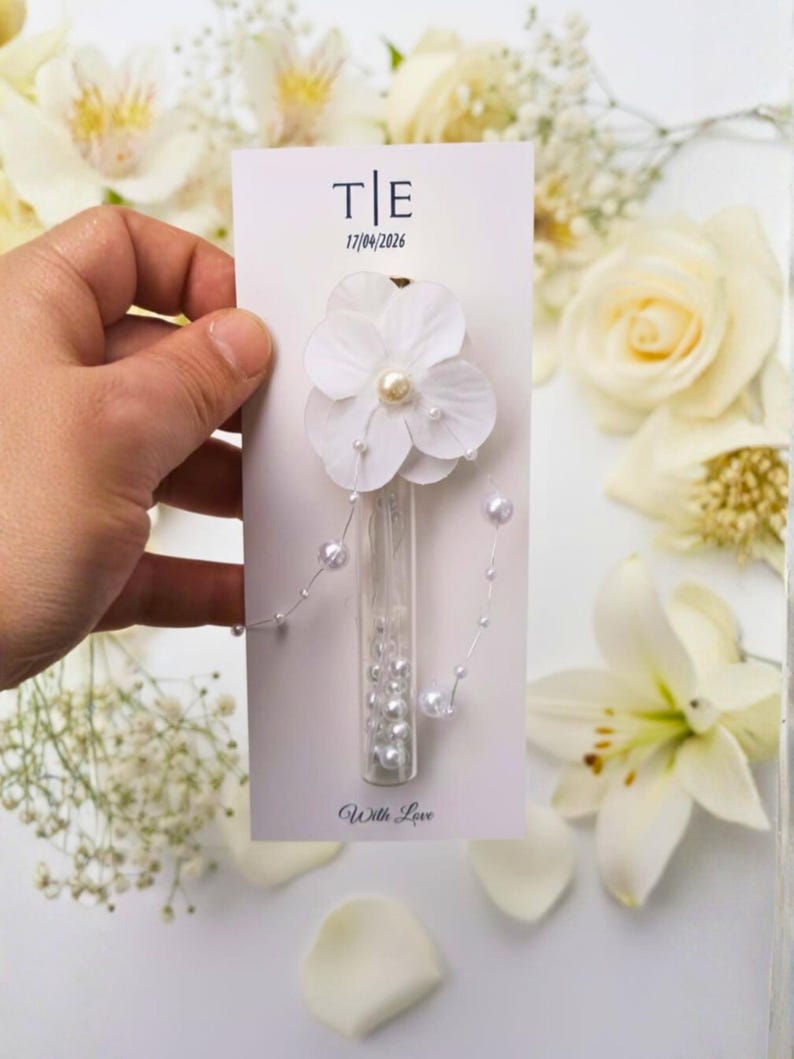 May include: A white floral wedding favor card with the initials "T|E" and the date "17/04/2026". The card features a white flower with a pearl center, a glass vial filled with pearls, and the words "With Love".