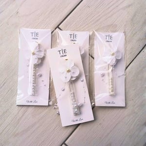 May include: Four wedding favors are displayed, each featuring a white flower with a pearl center and a clear tube filled with pearls. The favors are packaged on white cards with the text "T|E 17/04/2026" and "With Love".