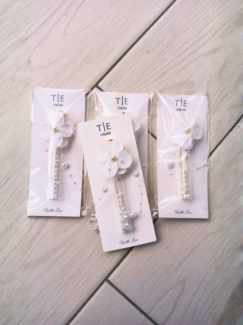 May include: Four wedding favors are displayed, each featuring a white flower with a pearl center and a clear tube filled with pearls. The favors are packaged on white cards with the text "T|E 17/04/2026" and "With Love".