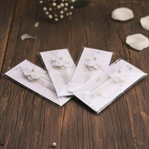 May include: Four white floral hair accessories with pearl accents are displayed on a dark wood surface. Each accessory is attached to a clear plastic tube and mounted on a white card. The word "TIE" is printed on the top left corner of each card.