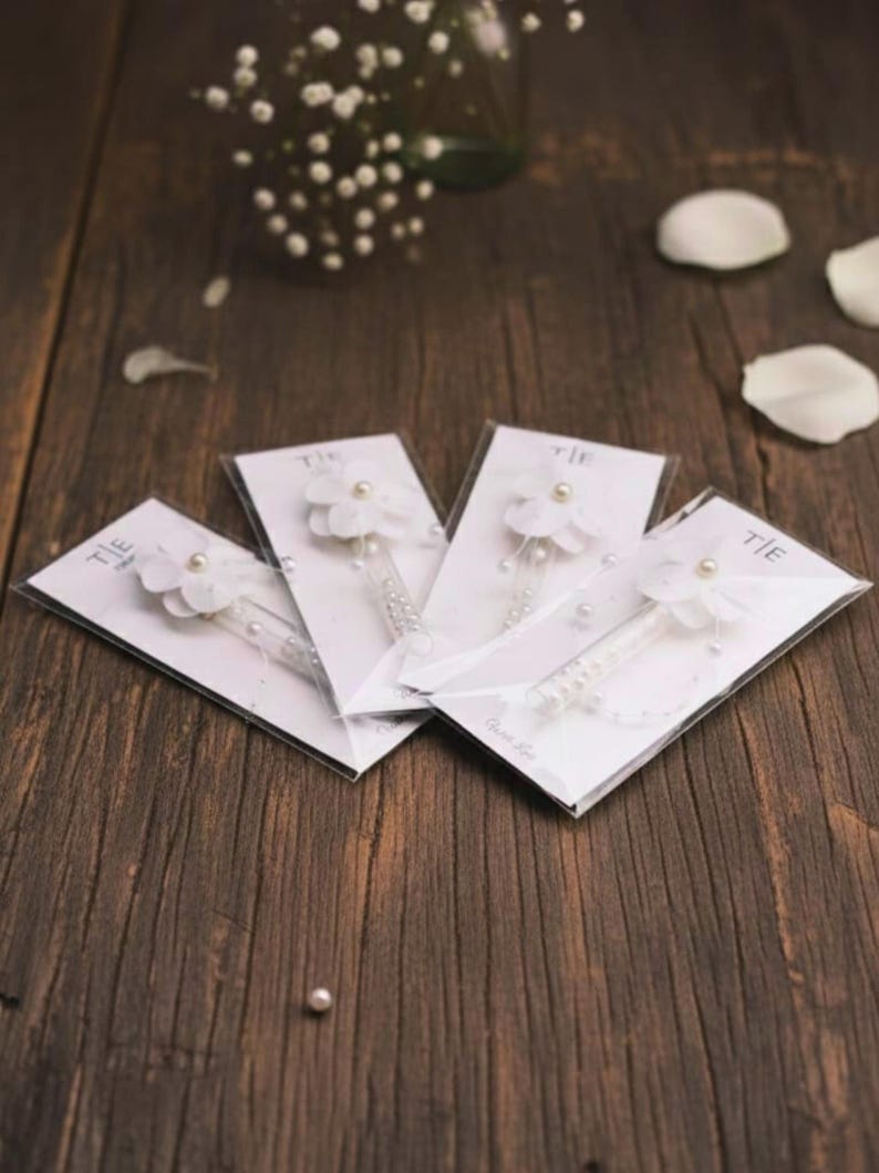 May include: Four white floral hair accessories with pearl accents are displayed on a dark wood surface. Each accessory is attached to a clear plastic tube and mounted on a white card. The word "TIE" is printed on the top left corner of each card.