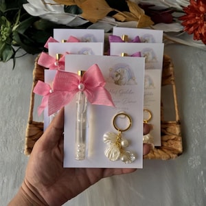 Op de afbeelding: Feestartikelen met bellenblaasstokken met roze strikken en parelaccenten, en sleutelhangers met parel- en bloemblaadjesbedels. De gunsten zijn gerangschikt op witte kaarten met een cartoonillustratie en tekst.