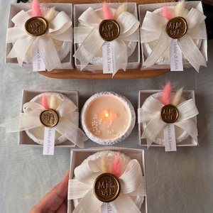 Op de afbeelding: Een verzameling witte, ronde decoratieve items, mogelijk zeep of kaarsen, gepresenteerd in kleine dozen. Elke doos is versierd met een wit lint, een gouden zegel en een label met de tekst "Melike & Eren".