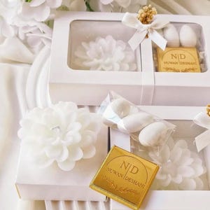 May include: White rectangular gift boxes containing a white flower-shaped candle, a small clear bag of white almond candies, and a square gold chocolate bar with the text N|D NUWAN DESHANI printed on it, accented with a white ribbon bow.