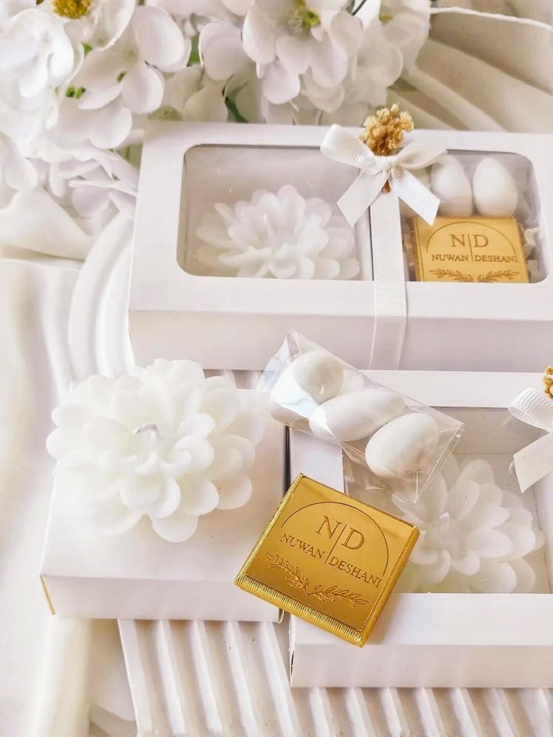 May include: White rectangular gift boxes containing a white flower-shaped candle, a small clear bag of white almond candies, and a square gold chocolate bar with the text N|D NUWAN DESHANI printed on it, accented with a white ribbon bow.