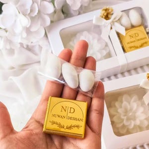 May include: A square gold-colored chocolate bar wrapper with the text ND NUWAN DESHANI and a leaf graphic, held alongside a clear plastic bag containing three white almond candies, with white gift boxes featuring similar branding in the background.