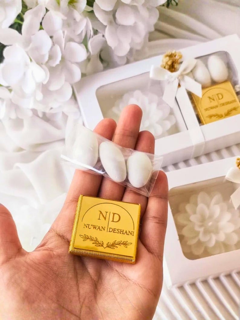 May include: A square gold-colored chocolate bar wrapper with the text ND NUWAN DESHANI and a leaf graphic, held alongside a clear plastic bag containing three white almond candies, with white gift boxes featuring similar branding in the background.