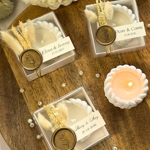 May include: Three square clear boxes containing white braided candle holders, each adorned with dried beige grasses and a gold wax seal featuring a leaf design. Each box includes a personalised tag with names and dates, plus one lit candle.