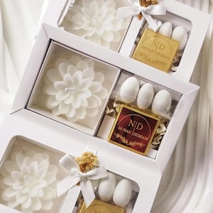 May include: Three white rectangular gift boxes, each containing a white flower-shaped candle, a small square plaque with the text ND NUWAN DESHANI, and white oval candies. The boxes are decorated with white ribbons and small dried floral accents.
