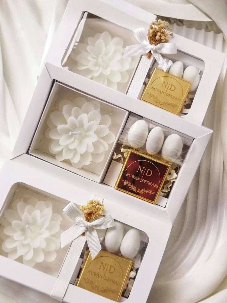 May include: Three white rectangular gift boxes, each containing a white flower-shaped candle, a small square plaque with the text ND NUWAN DESHANI, and white oval candies. The boxes are decorated with white ribbons and small dried floral accents.
