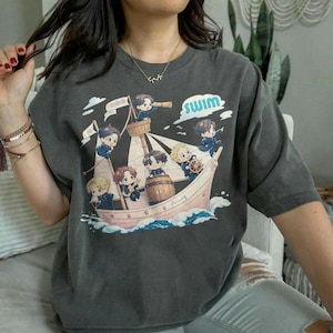 Puede incluir: Camiseta gris oscuro con una ilustración de dibujos animados de un barco con personajes animados. El barco es rosa y blanco, con la palabra "swim" en azul y blanco. Los personajes llevan uniformes azules.