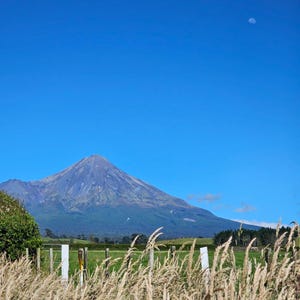May include: A scenic landscape featuring a mountain under a clear blue sky with a crescent moon. Tall, dry grass sways in the foreground, with a fence and green fields. The mountain has a dark, rocky appearance.
