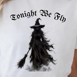 Könnte beinhalten: Ein weißes T-Shirt mit einer schwarzen grafischen Illustration einer Hexe, die einen spitzen Hut trägt und einen Besen hält. Über der Figur ist der Text Tonight We Fly in einer schwarzen, gotischen Schriftart mittig aufgedruckt.