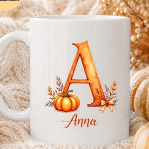 Könnte beinhalten: Ein weißer Keramikbecher mit einem großen orangefarbenen Buchstaben A, der mit einem kleinen Kürbis, Herbstblättern und einer Schleife verziert ist. Der Name Anna ist in orangefarbener Schreibschrift unter dem Buchstaben gedruckt.