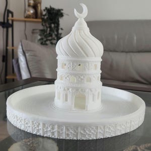 May include: A white, 3D-printed mosque model with a crescent moon finial. The mosque has detailed architectural features and sits on a round, decorative base. The model is displayed on a glass table.