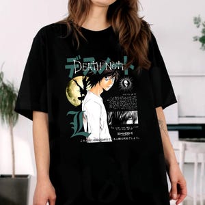 May include: Black t-shirt featuring anime artwork with the text "Death Note" in English and Japanese. The design includes a character illustration, a moon, and additional Japanese text. The shirt is a casual wear item.