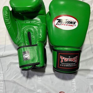 Könnte beinhalten: Paar leuchtend grüne Boxhandschuhe. Einer der Handschuhe zeigt ein Twins Special-Logo in einem roten Oval. Der andere Handschuh hat ein Twins Special-Logo am Handgelenkriemen. Die Handschuhe sind aus glänzendem Material.