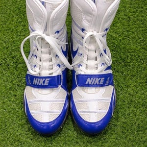 May include: Pair of white and blue Nike wrestling shoes. The shoes have a high-top design with white laces, a blue strap with the Nike logo, and blue accents. The shoes are on a green surface.