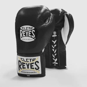 Könnte beinhalten: Schwarze Leder-Boxhandschuhe mit weißen Schnürsenkeln. Die Handschuhe tragen das Logo "CLETO REYES" in Weiß und Gold. Profi-Boxhandschuhe, hergestellt in Mexiko.