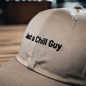 Può includere: Un cappellino da baseball beige con la scritta ricamata in nero "Just a Chill Guy". Il cappellino ha una visiera curva e un bottone sulla parte superiore. Il tessuto sembra essere cotone resistente o simile.
