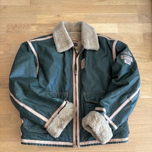 Può includere: Una giacca verde scuro con colletto e polsini in pelliccia sintetica beige. La giacca presenta una chiusura frontale con zip, due tasche frontali con patta e profili beige lungo le cuciture. Una toppa sulla manica sinistra riporta la scritta N.Y.W. VICTORY DIESEL 557.
